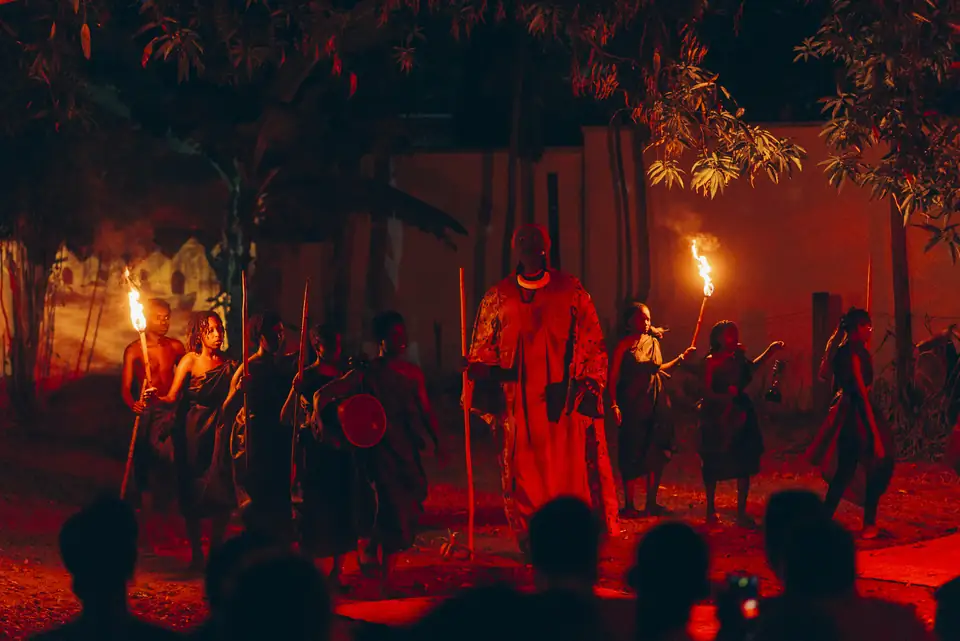 Inamujandi, juillet 2025 : une guerrière en robe rouge entourée de porteurs de torches, devant le public, dans le jardin du centre